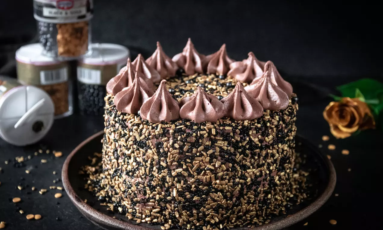 Nyårstårta täckt med svart och guldigt strössel, toppad med bruna chokladrosetter på en mörkbrun tallrik mot svart bakgrund