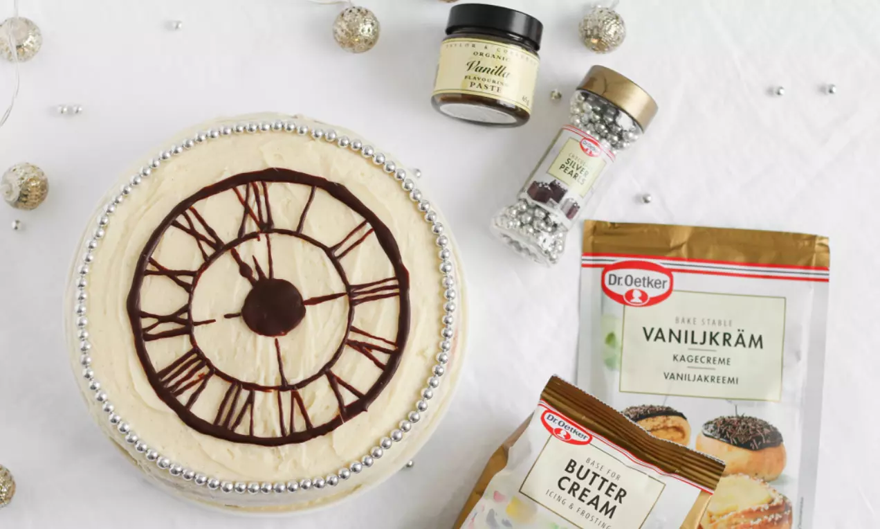 Nyårstårta med vaniljkräm och chokladklocka på ljus botten, dekorerad med silvriga pärlor, omgiven av bakingredienser på vit duk