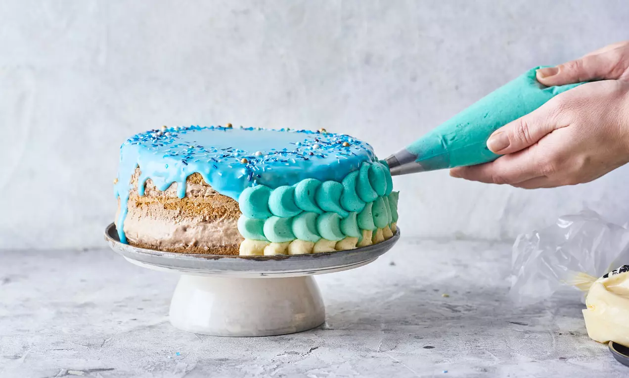 Mermaidtårta med blå glasyr, dekorerad med spritsade turkosa och vita vågmönster, stående på vit tårtställning mot ljus bakgrund