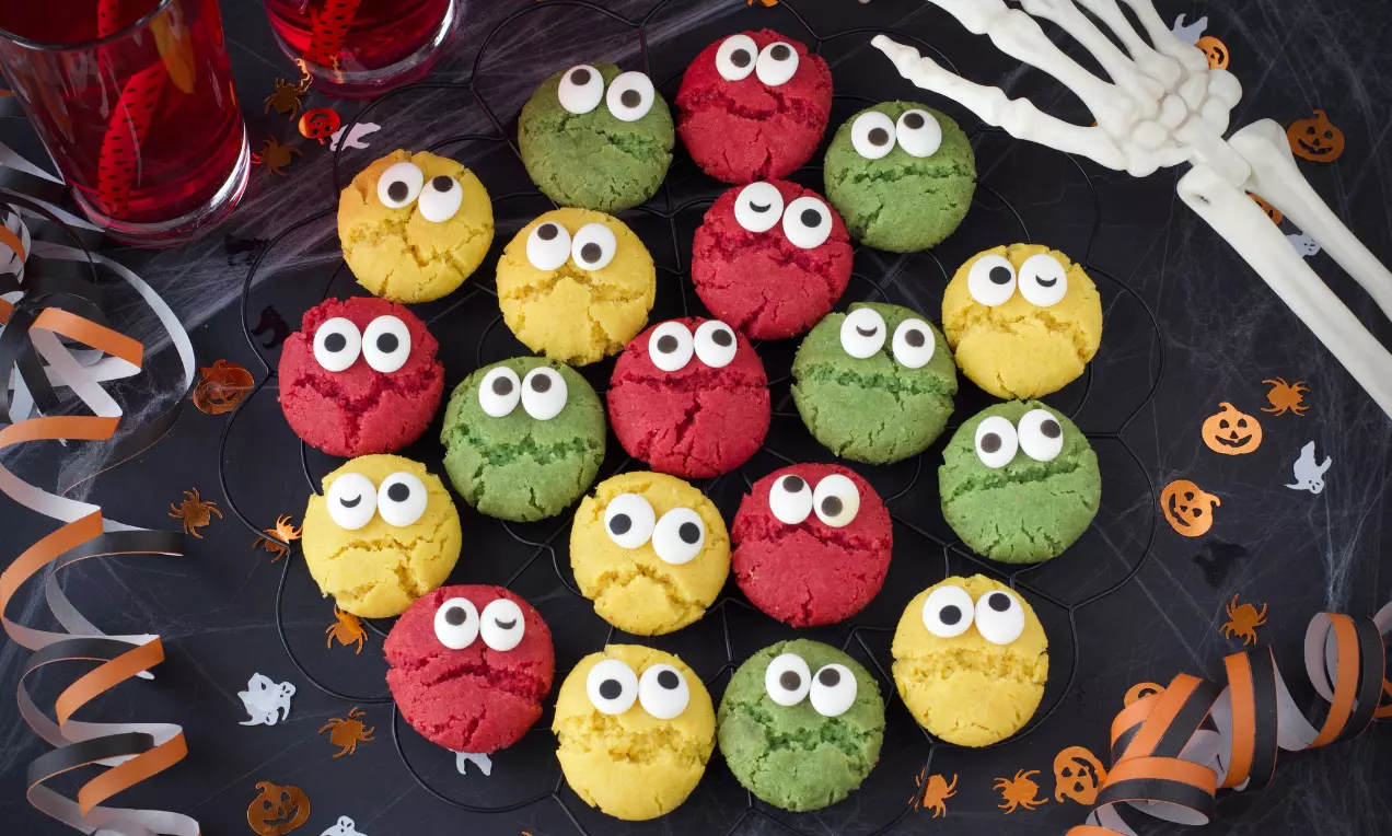 Färgglada småkakor i gul, röd och grön färg med sprucken yta och vita ögon på svart fat med Halloween-dekorationer som bakgrund