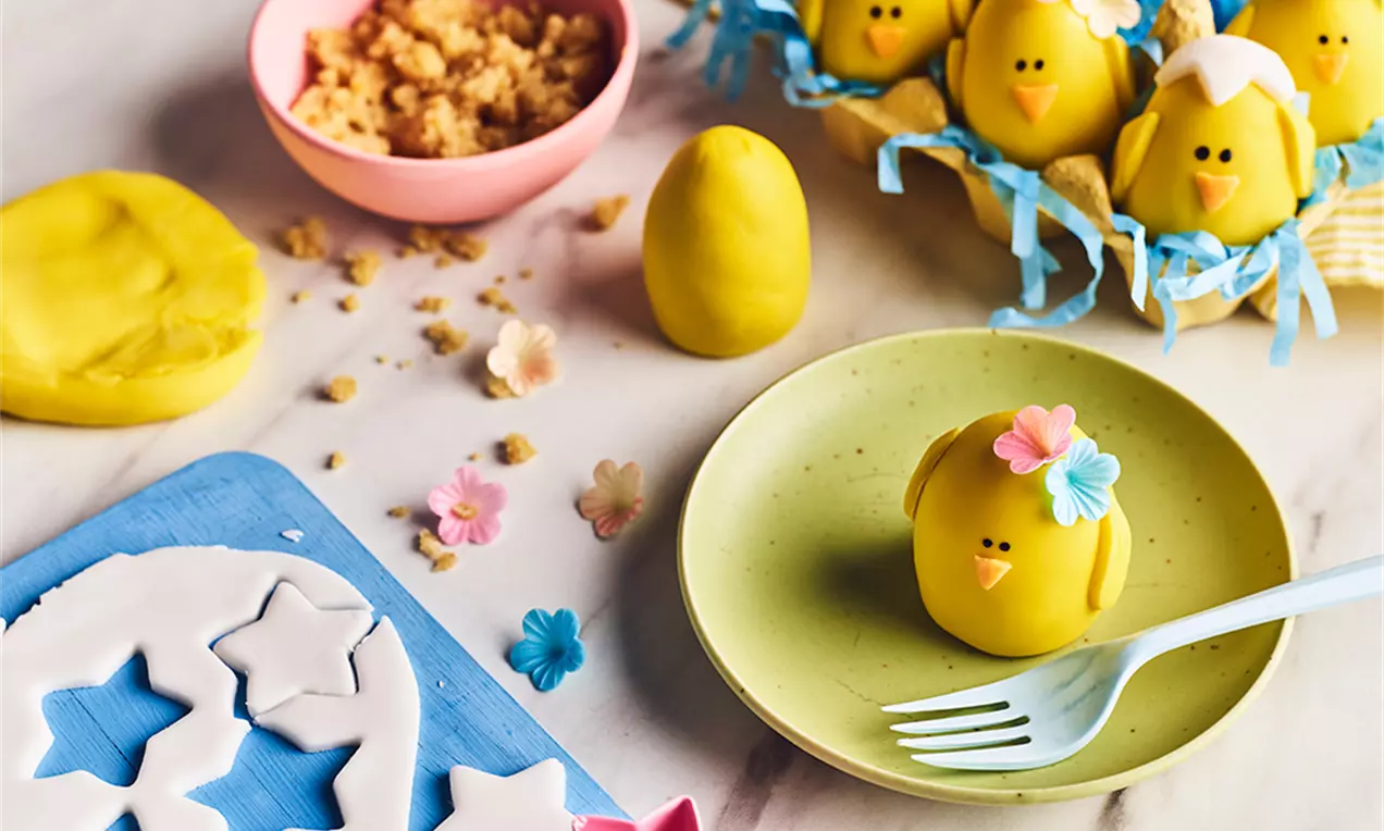 Gula marsipankycklingar med vita detaljer och blommor, formade som ägg på ett blått fat med pappersstrimlor och marsipanpyssel