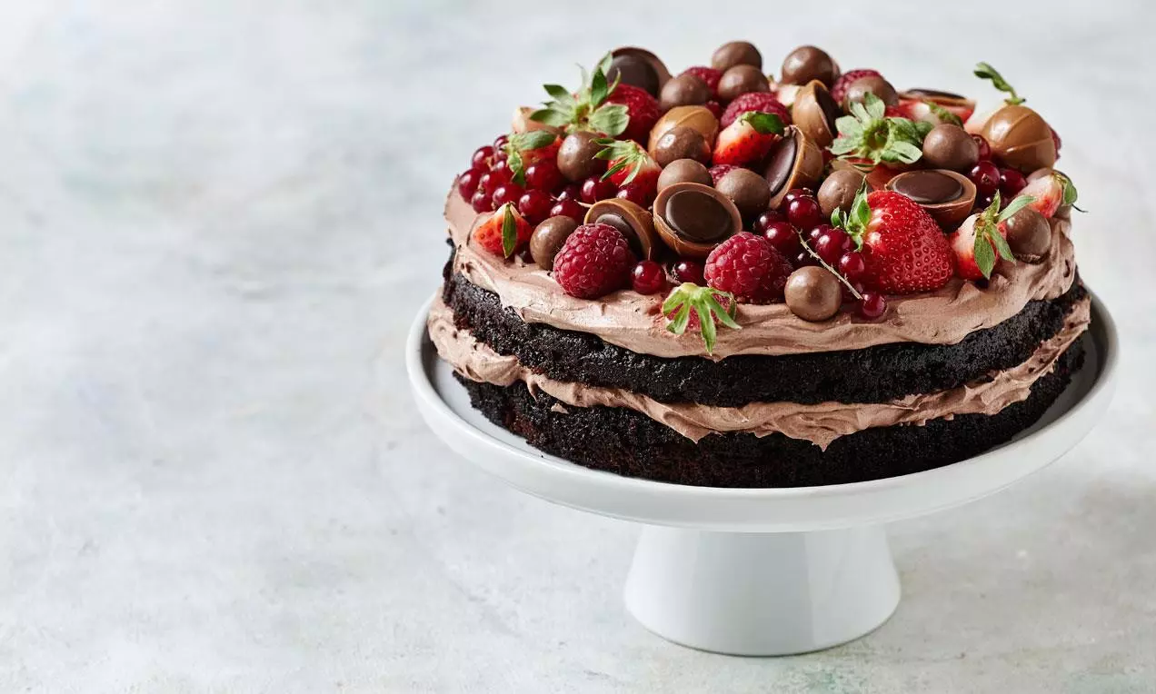 Chokladtårta med tjocka lager av brun chokladkräm, toppad med färska bär, chokladkulor och praliner på vitt fat med stjälkar