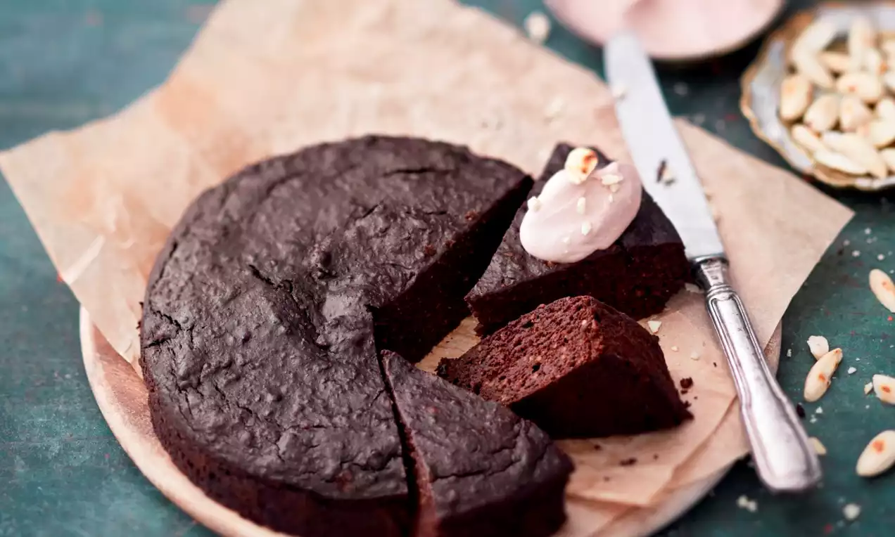 Rund chokladkaka på brunt bakpapper med utskurna bitar, en toppad med ljusrosa kräm bredvid kniv, nötter och skål på grön bakgrund