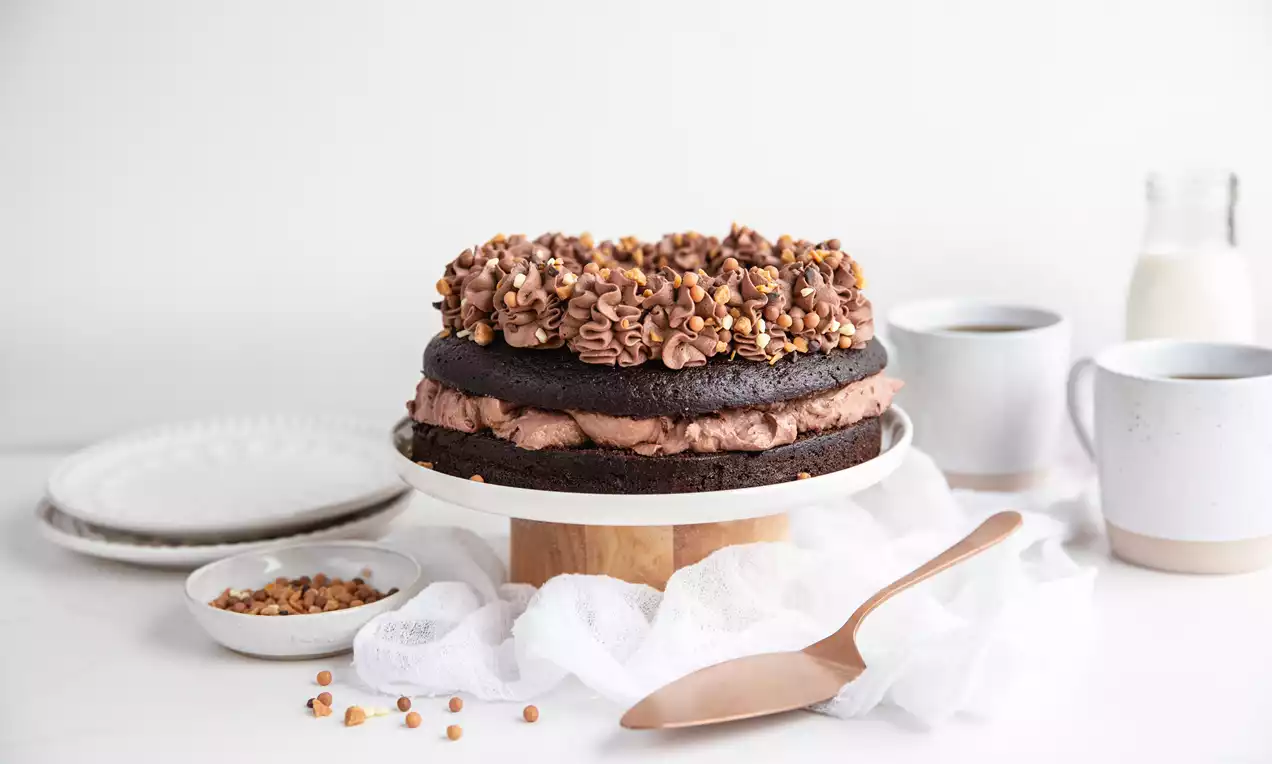 Mörk chokladkaka i två lager med krämig fyllning, toppad med spritsad chokladkräm och strödd med gyllenbruna nötter på ett vitt kakfat