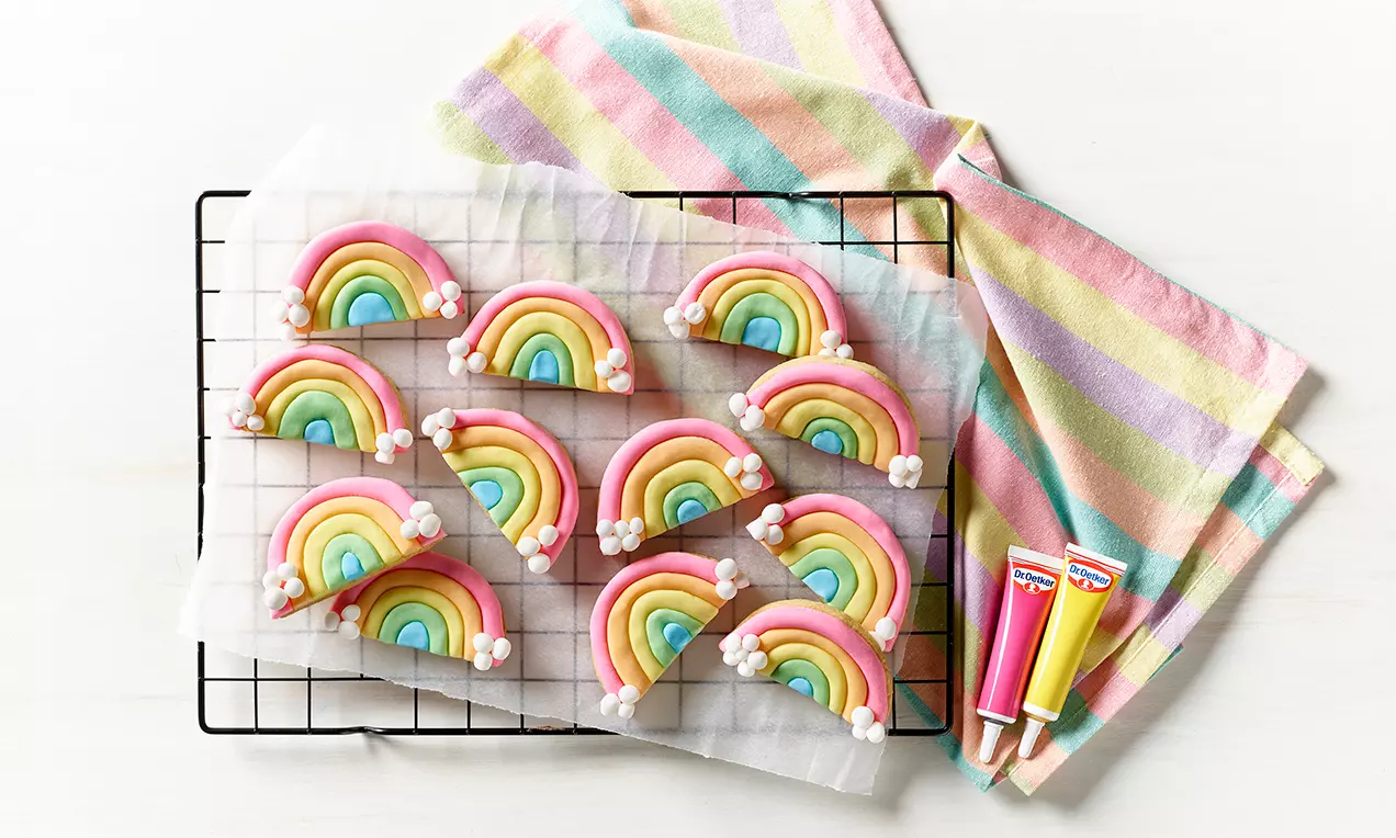 Halvmåneformade regnbågskakor i rosa, gult, grönt och blått med vita sockersmå moln på ett galler med bakplåtspapper och randig handduk