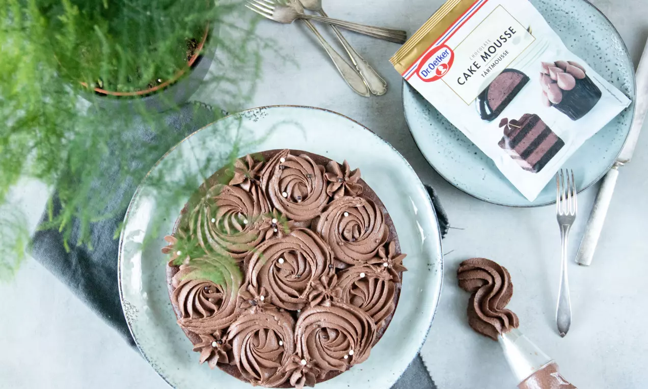 Rund kladdkaka täckt med virvlande chokladmousse, toppad med strössel på en ljusgrå tallrik, omgiven av gröna blad och bakredskap
