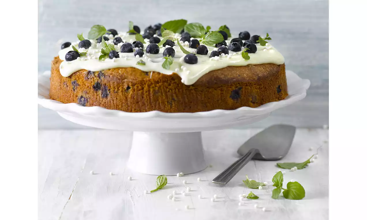 Blåbärskaka med guldbrun yta, vit frosting toppad med blåbär och gröna blad, placerad på vit tårtfat med silverspatel bredvid