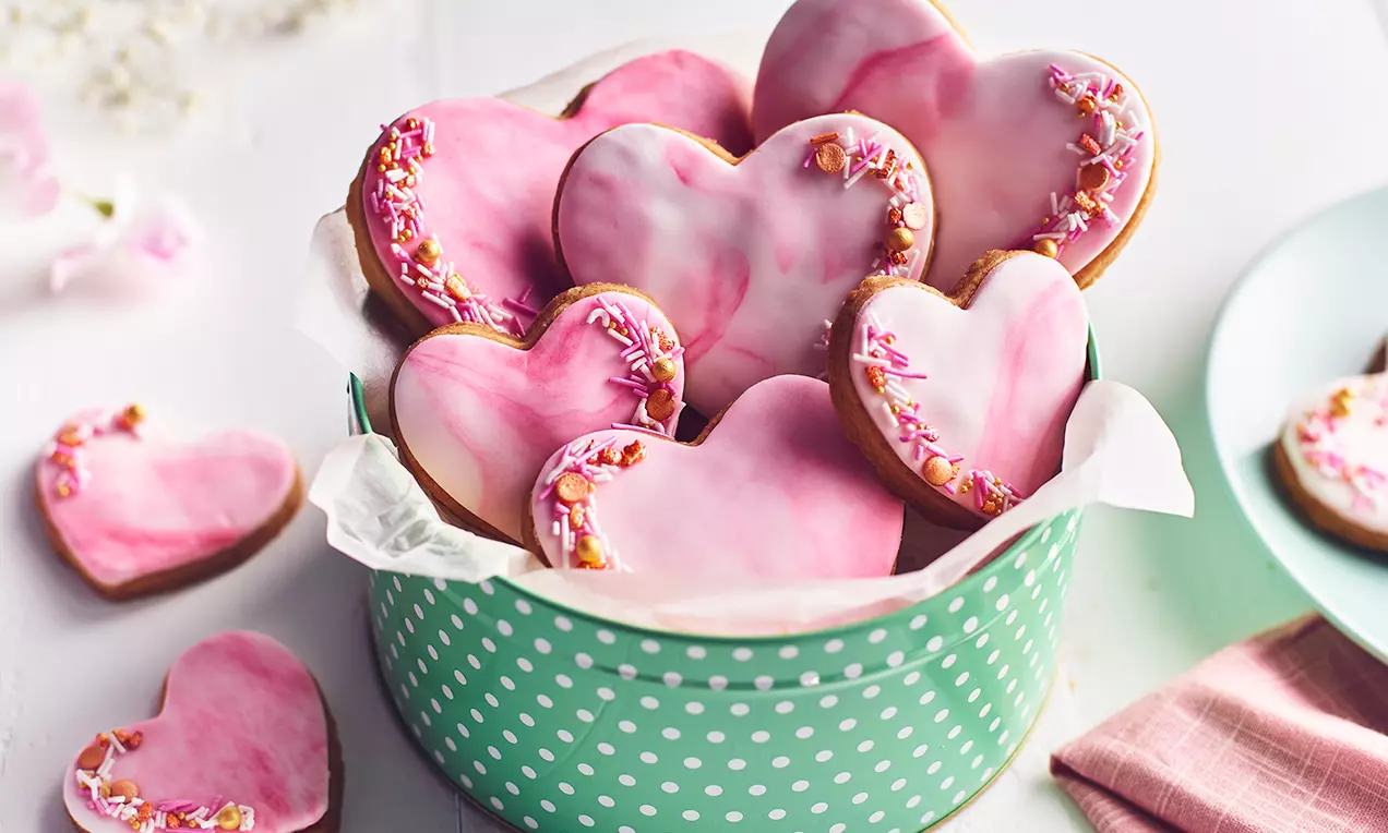 Hjärtkakor med rosa och vit marmorerad glasyr, dekorerade med strössel längs kanten, lagda i en grön prickig ask med bakplåtspapper