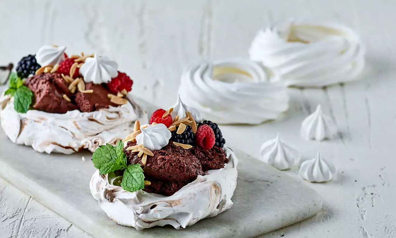 Marängpavlova med vit spröd bas, toppad med chokladkräm, färska bär, mandelspån, marängtoppar och gröna myntablad på ljus stenbricka