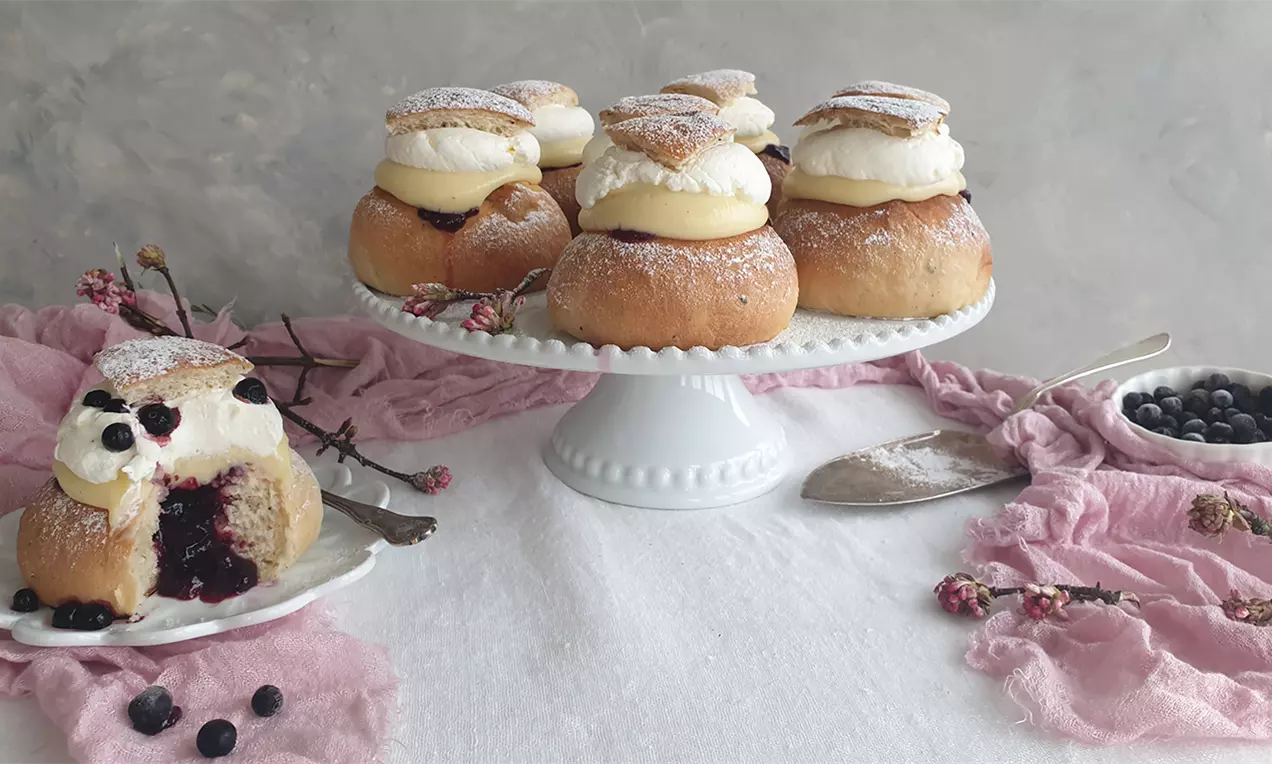 Vaniljsemlor med gyllene bullar, vispad grädde och florsocker, fyllning av vaniljkräm och blåbär på vit kakbricka med rosa tyg och bärskål