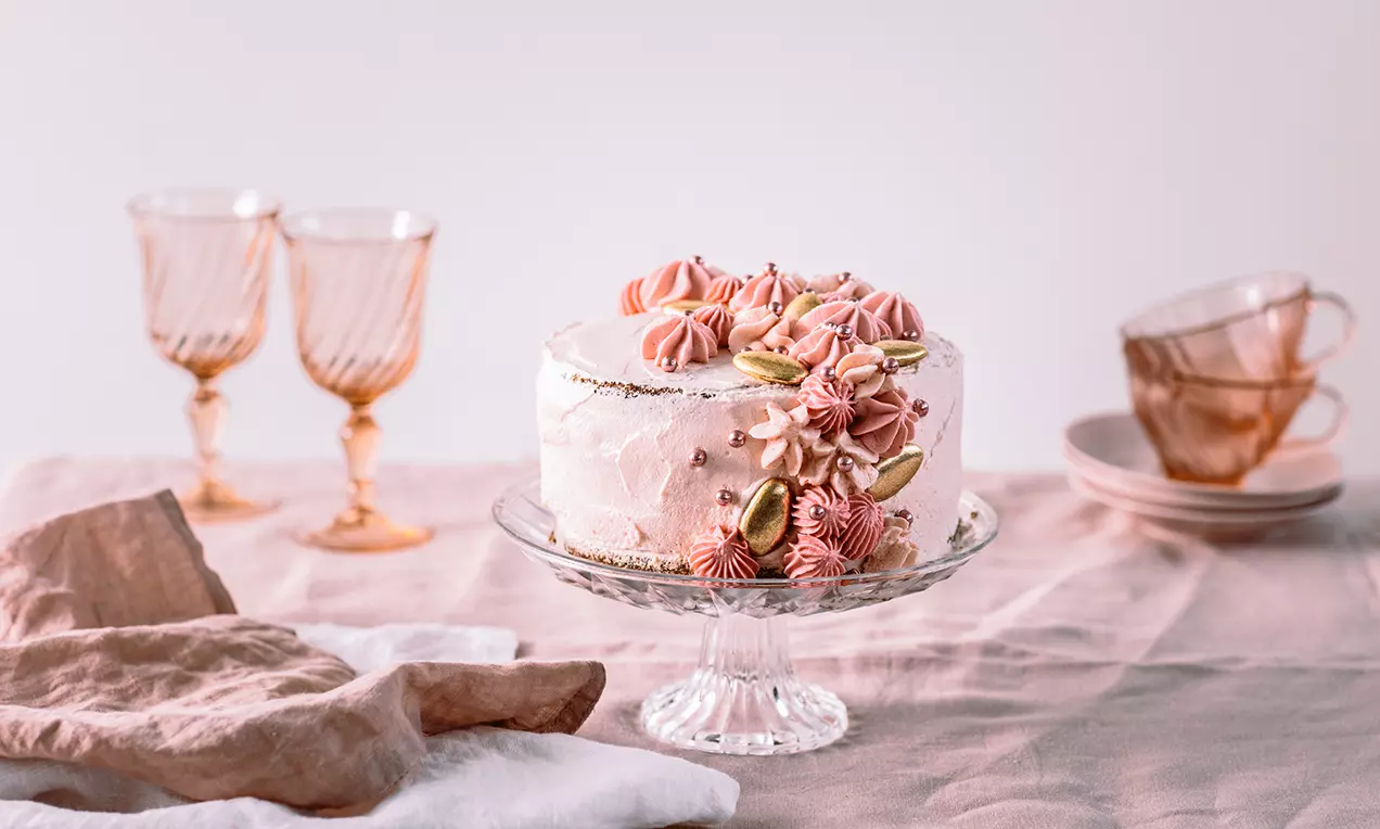 Rose & Gold-tårta med rosatonade gräddrosor och gulddekorationer på glasfat, omgiven av rosa textilier och glasdetaljer