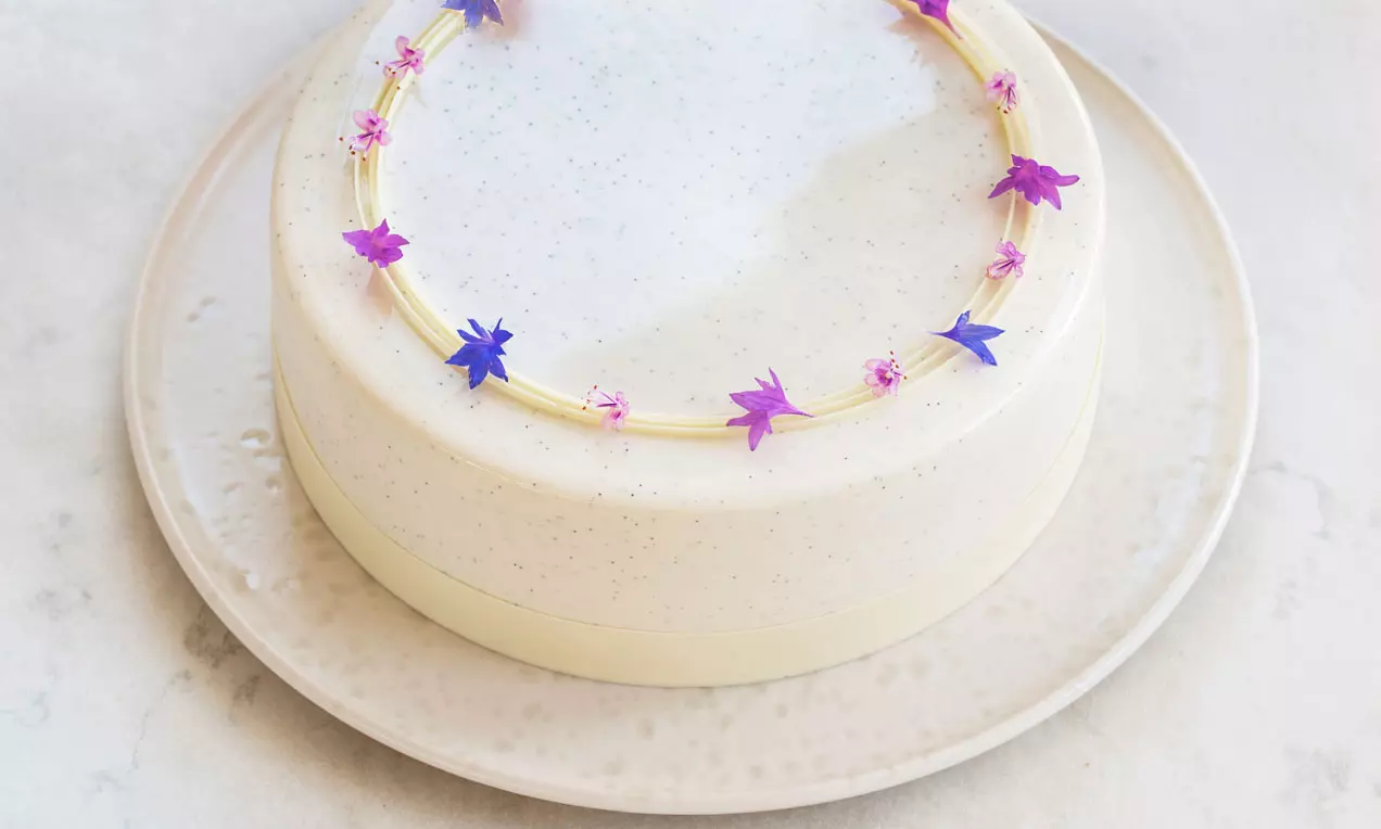 Vanilj- och blåbärsmoussetårta med glänsande vit vaniljglaze, dekorerad med små rosa och lila blommor på en ljus keramikfat