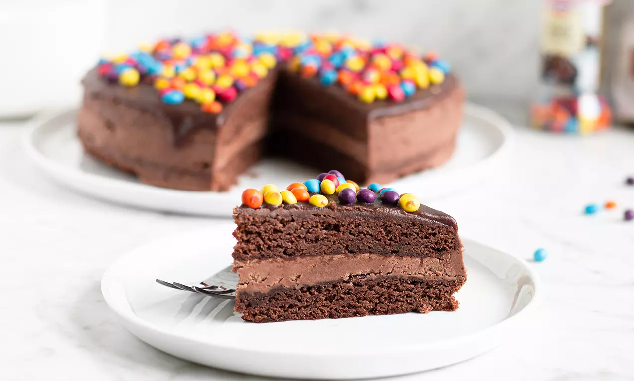 Kladdkaketårta med två lager chokladfyllning, dekorerad med färgglada chokladlinser, serverad med en tårtbit på vit tallrik