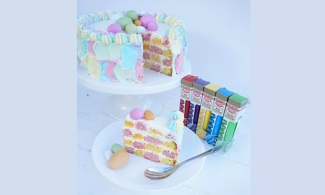 Påsktårta med lager av rosa och gula bottnar, vit glasyr, pastellfärgade frostingdekorationer och chokladägg på ett vitt fat