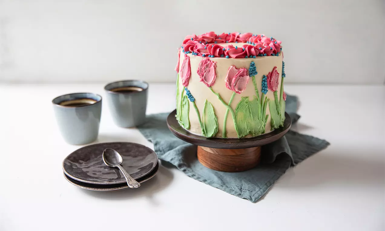 Tårta på träfat täckt med grön frosting och rosa tulpaner, toppad med rosor, omgiven av kaffekoppar och fat på grå linneservett