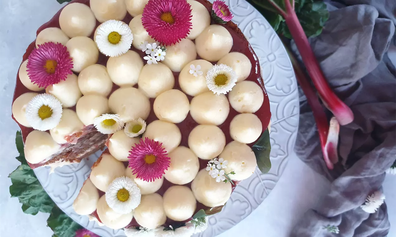 Morotstårta med röd rabarber- och kardemummapuré, toppad med ljusa krämkulor och små vita och rosa blommor på ett mönstrat tårtfat