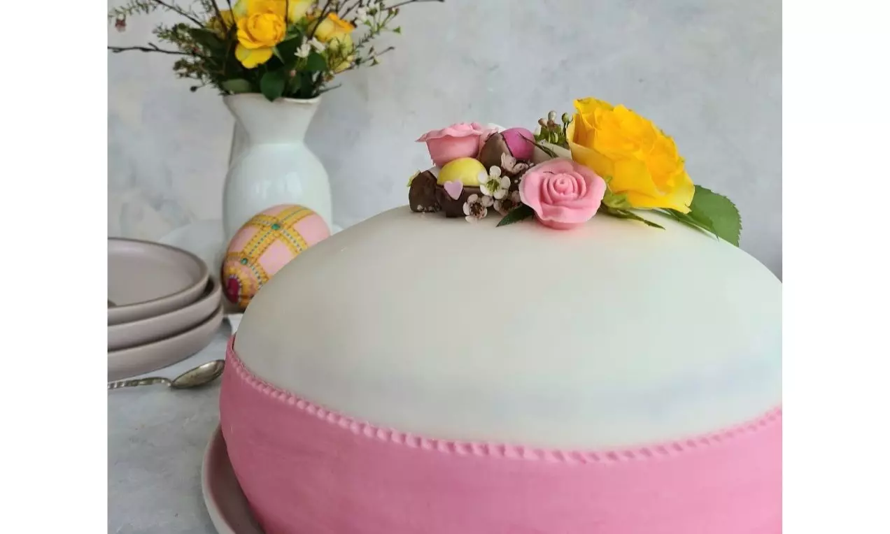 Rund gräddvit tårta med rosa marsipanband dekorerad med gula och rosa rosor, tårtans ljusa yta kontrasterar mot den pastellfärgade bakgrunden