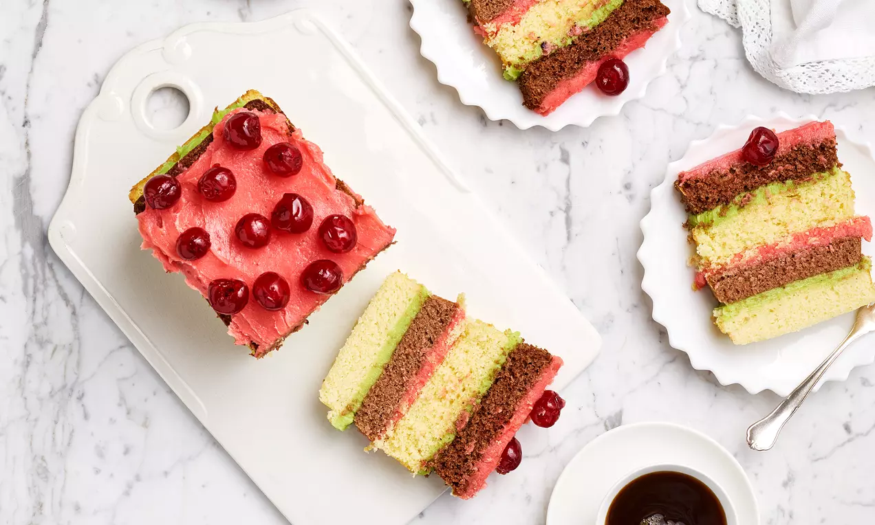 Randig tårta med lager av grön, gul och brun botten, täckt med rosa glasyr och röda körsbär, serverad på vit tårtbricka