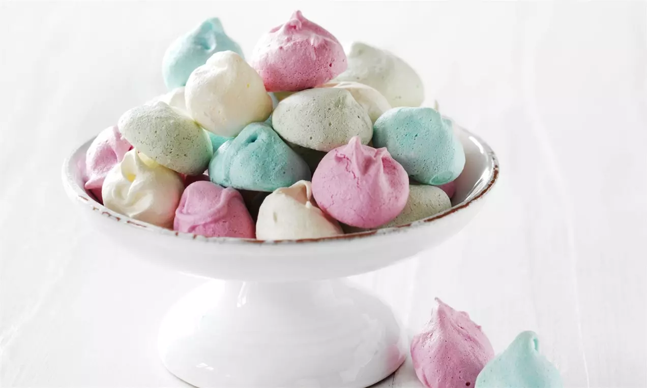 Pastellfärgade maränger i rosa, vitt, grönt och blått med krispig yta placerade på ett vitt fat med hög fot mot ljus bakgrund