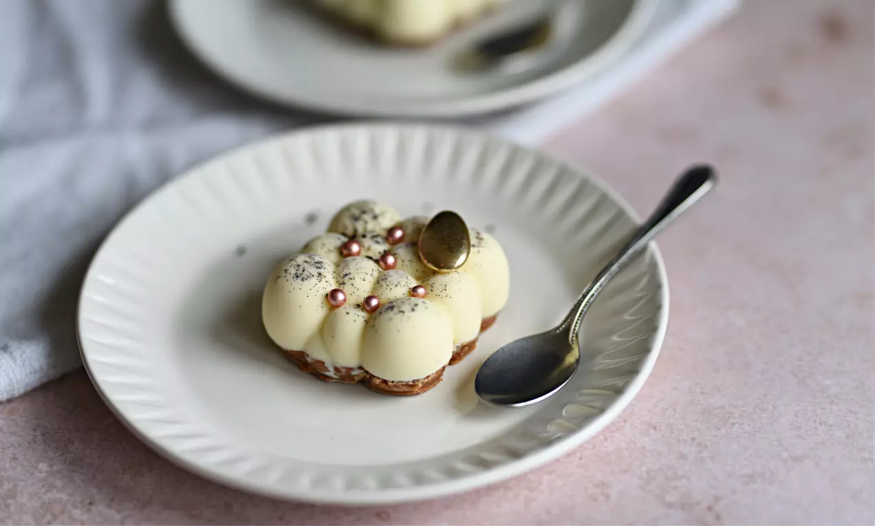 Gul vaniljmousse formad i sfäriska kupoler på brun chokladbotten, dekorerad med äppelbitar, pärlor och bladguld på vit tallrik