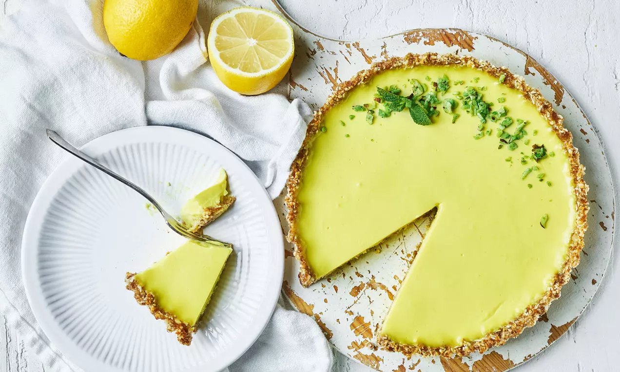 Vegansk citrontårta med ljusgul, slät fyllning i en brun nötbotten, toppad med gröna örter och serverad med citroner på ett rustikt bord